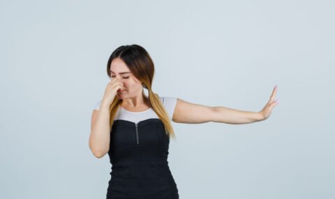 young blonde woman showing stop gesture while pinching nose in dress and looking disgusted , front view.