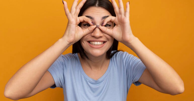 smiling-pretty-brunette-caucasian-girl-looks-camera-through-fingers-orange